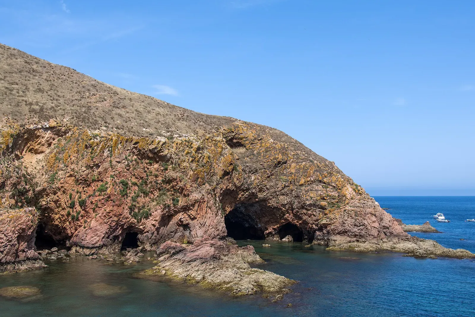 Sea caves cut into pink granite cliffs above clear blue water, with small boats offshore