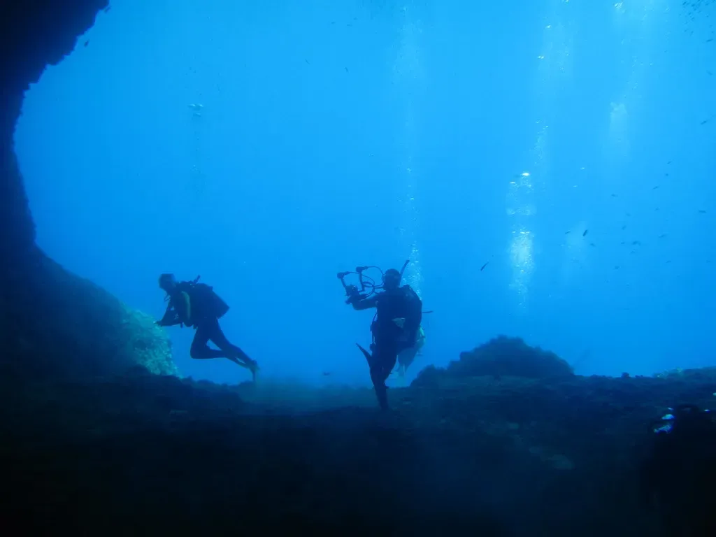 Two divers over rocky reef in clear blue water outside a cavern