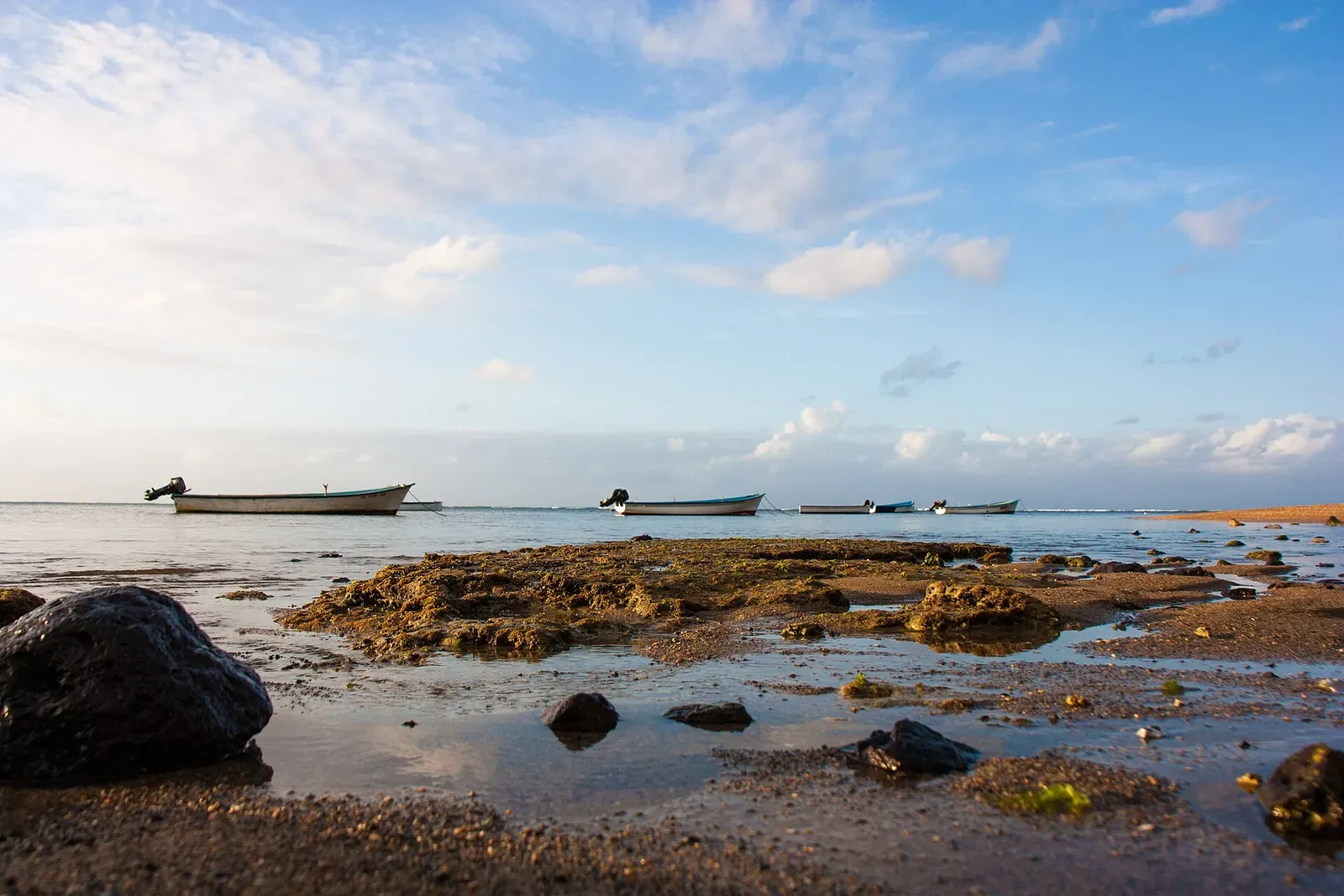 Small fishing boats moored in the shallow lagoon off Rodrigues at low tide, with exposed reef rock in the foreground.