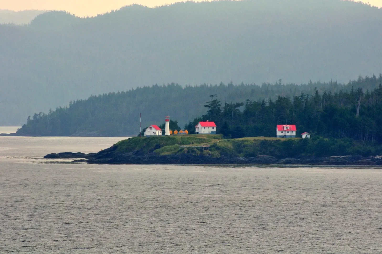A red-roofed lighthouse station sits on a forested point above the water near Port Hardy