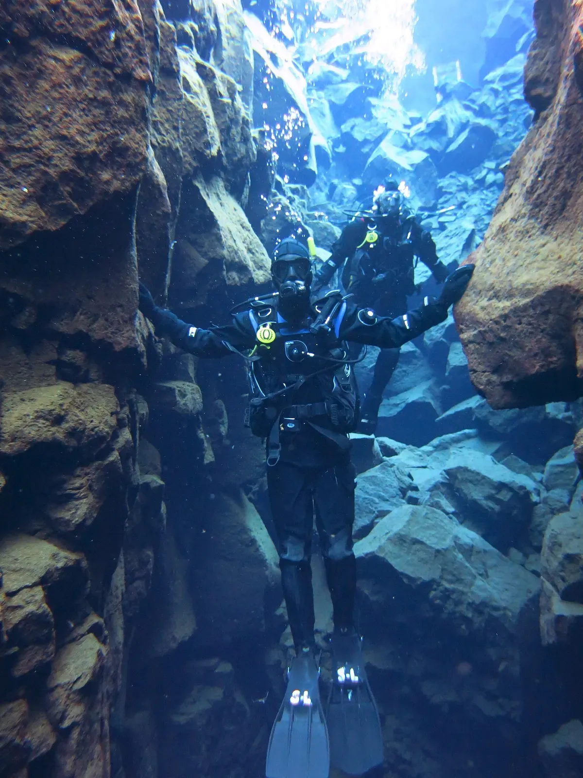 Two divers in drysuits float through a narrow basalt corridor in the Silfra fissure, lit by shafts of daylight