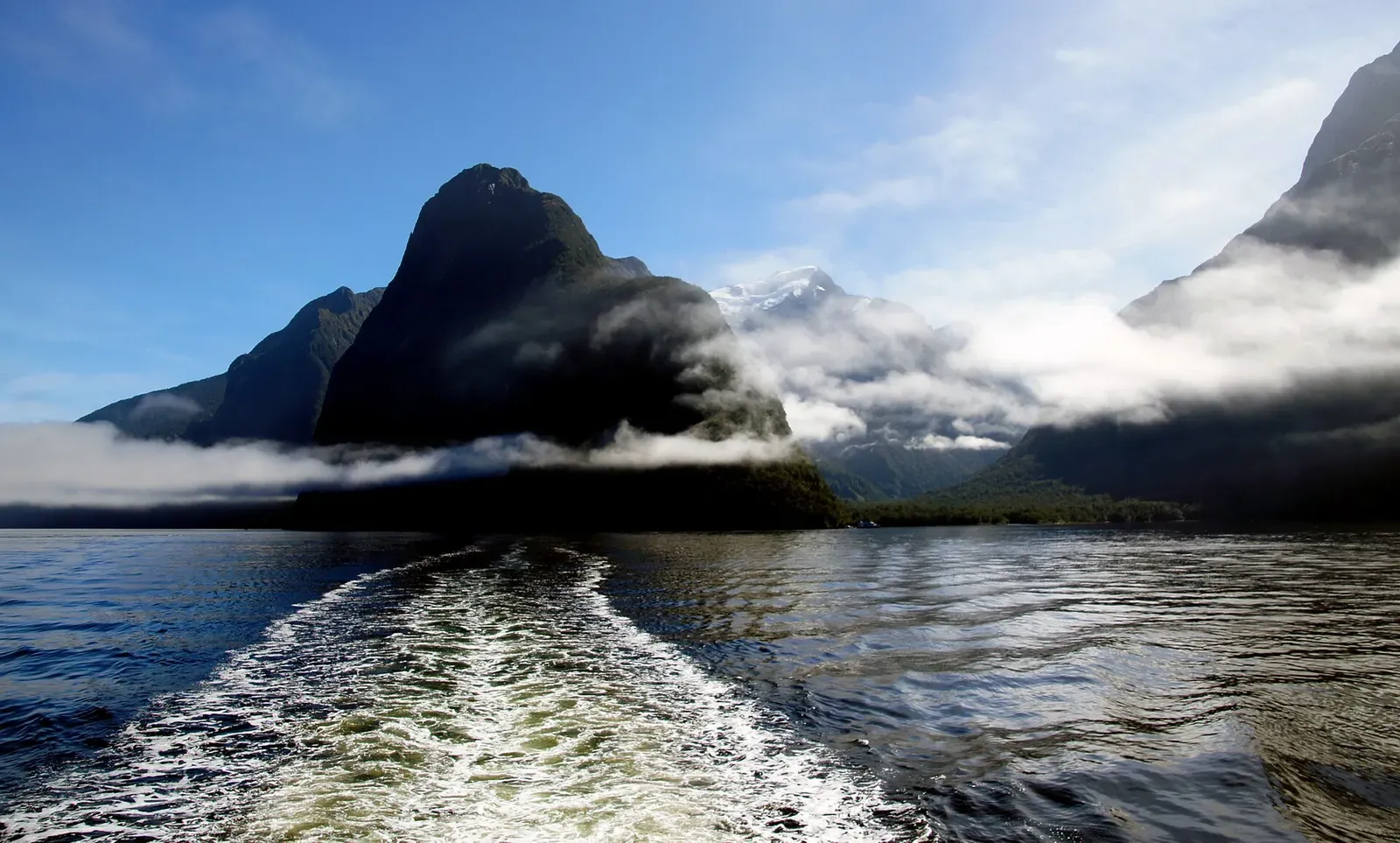 Boat wake crossing Milford Sound beneath mist-wrapped fjord peaks
