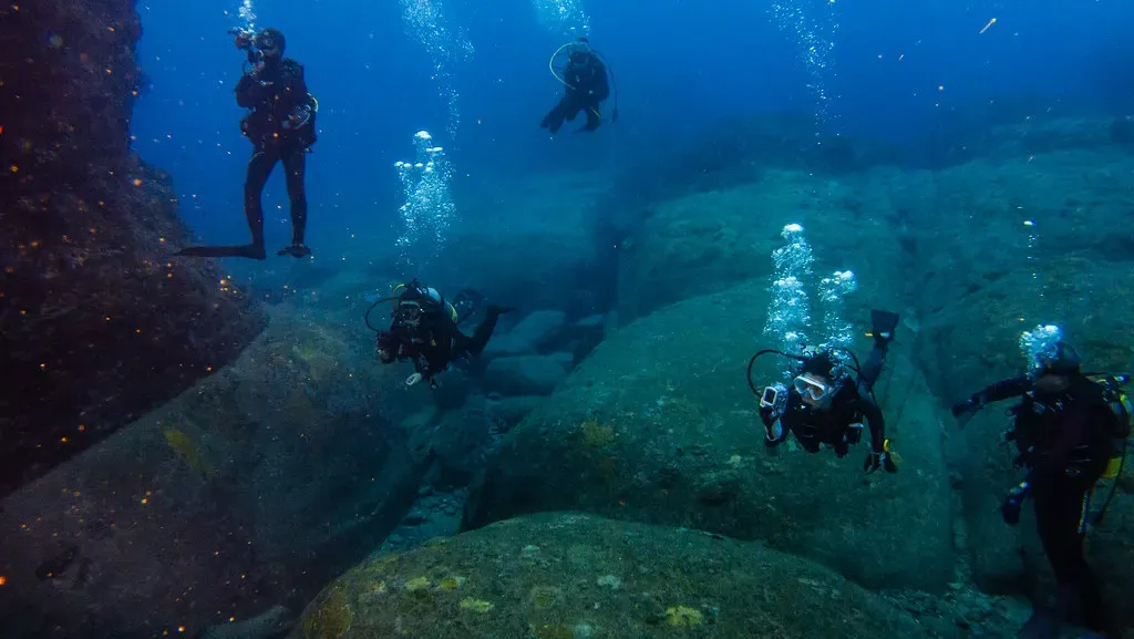 Divers hover among sunlit rock formations and rising bubble trails at a Yonaguni dive site.