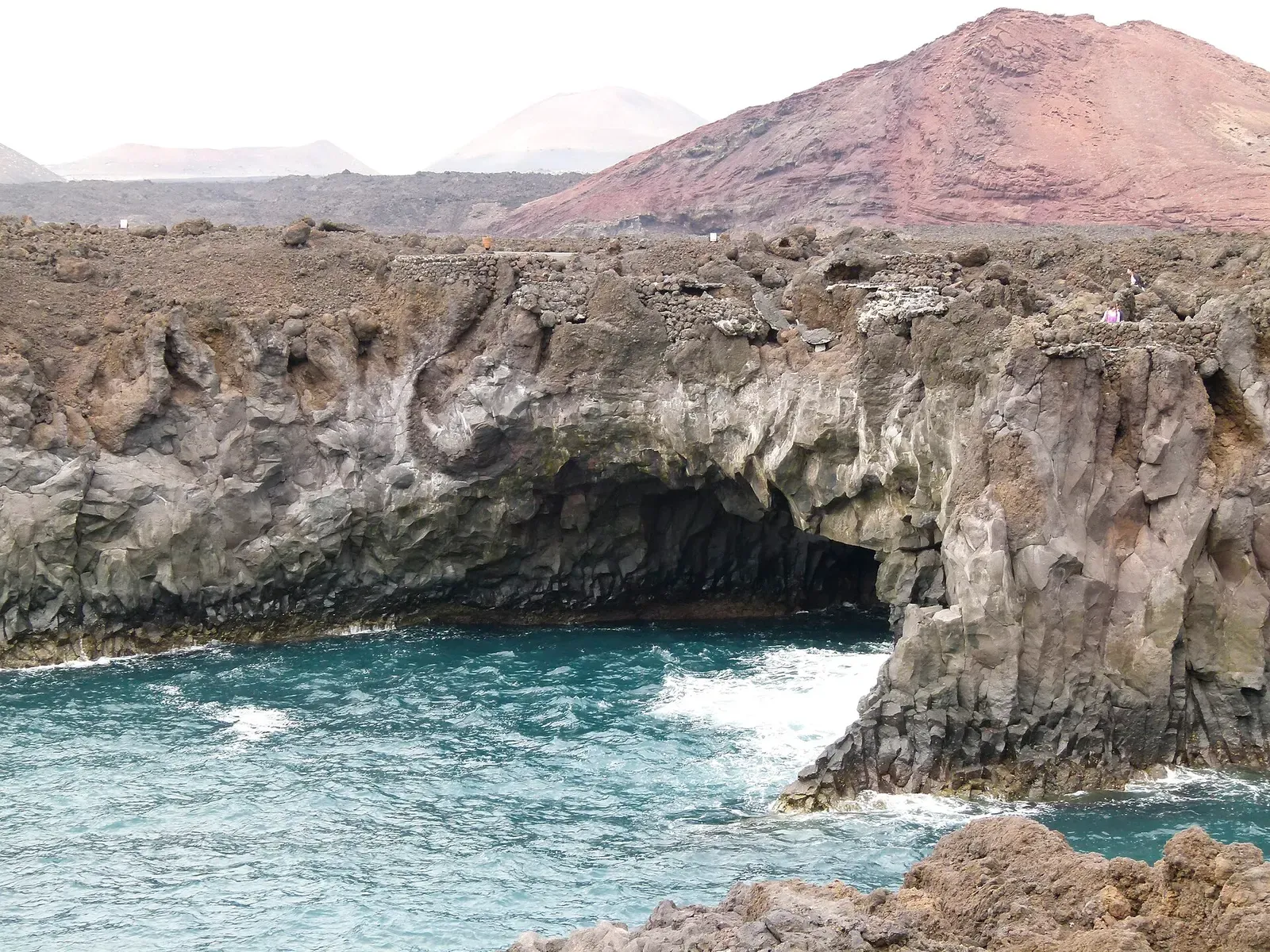 Waves surge into a lava sea arch below the red volcanic cone of Montaña Bermeja at Los Hervideros