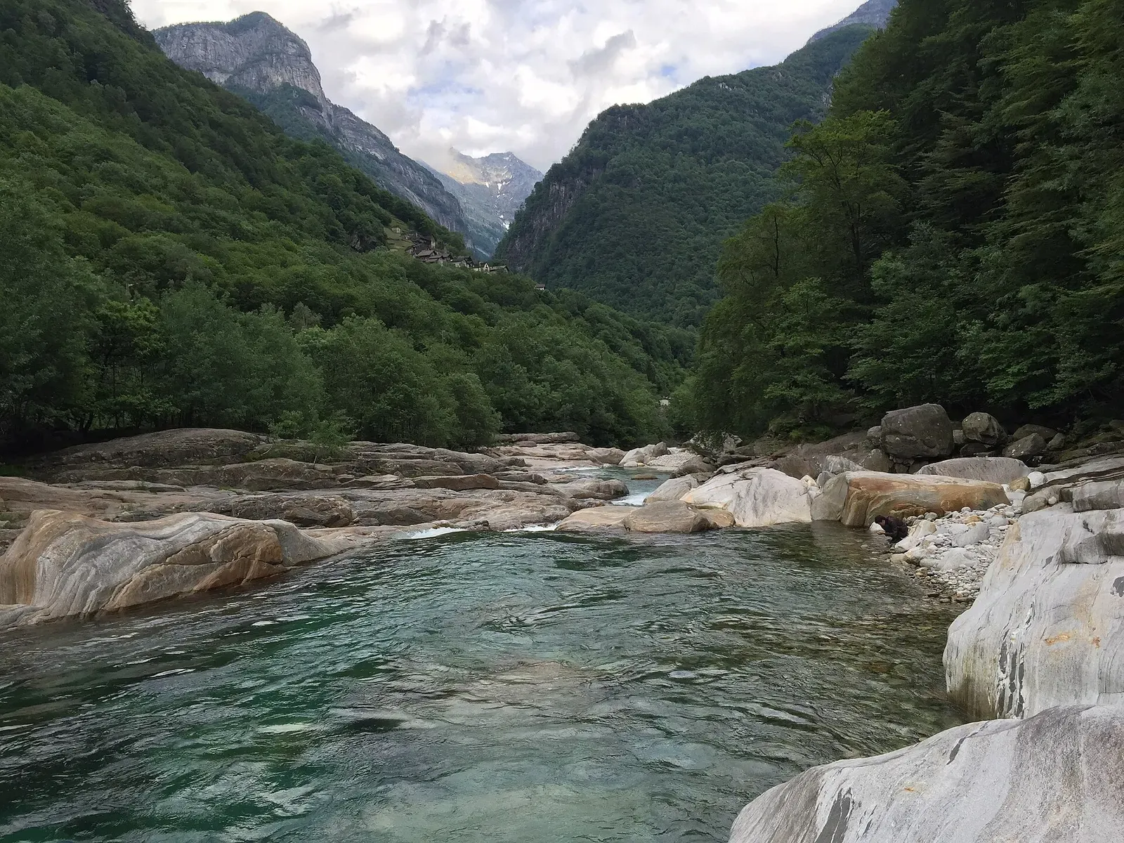 Verzasca valley view down a clear green pool toward forested mountains and snow-capped peaks