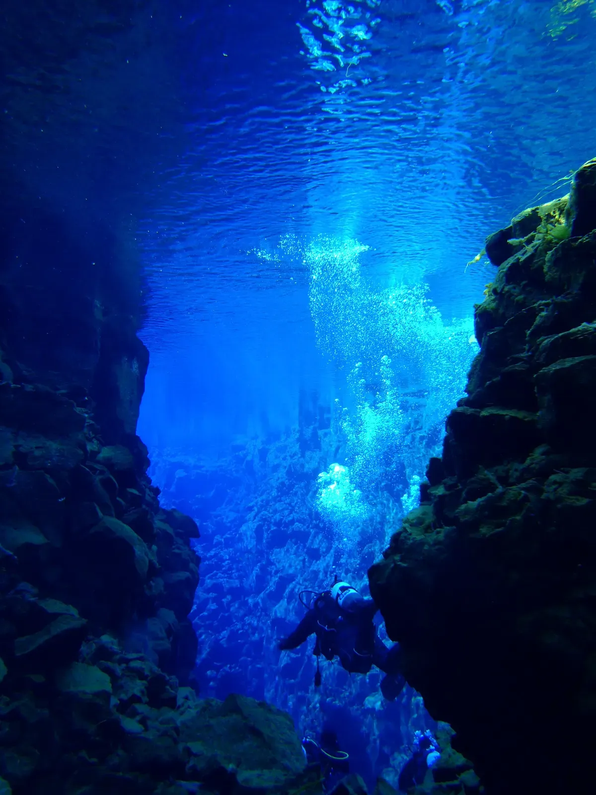 A scuba diver descends through a deep blue crevasse in Silfra with a trail of exhaled bubbles rising past the rock walls