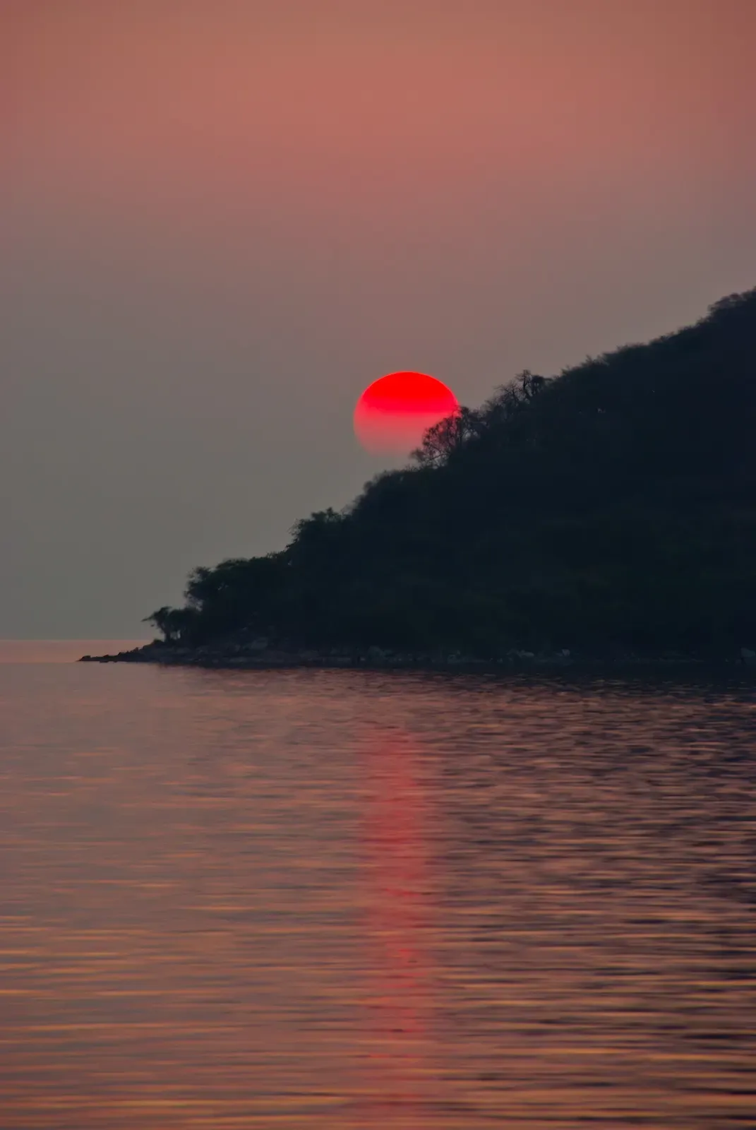 The sun sets a deep red behind a wooded headland on the shore of Lake Malawi