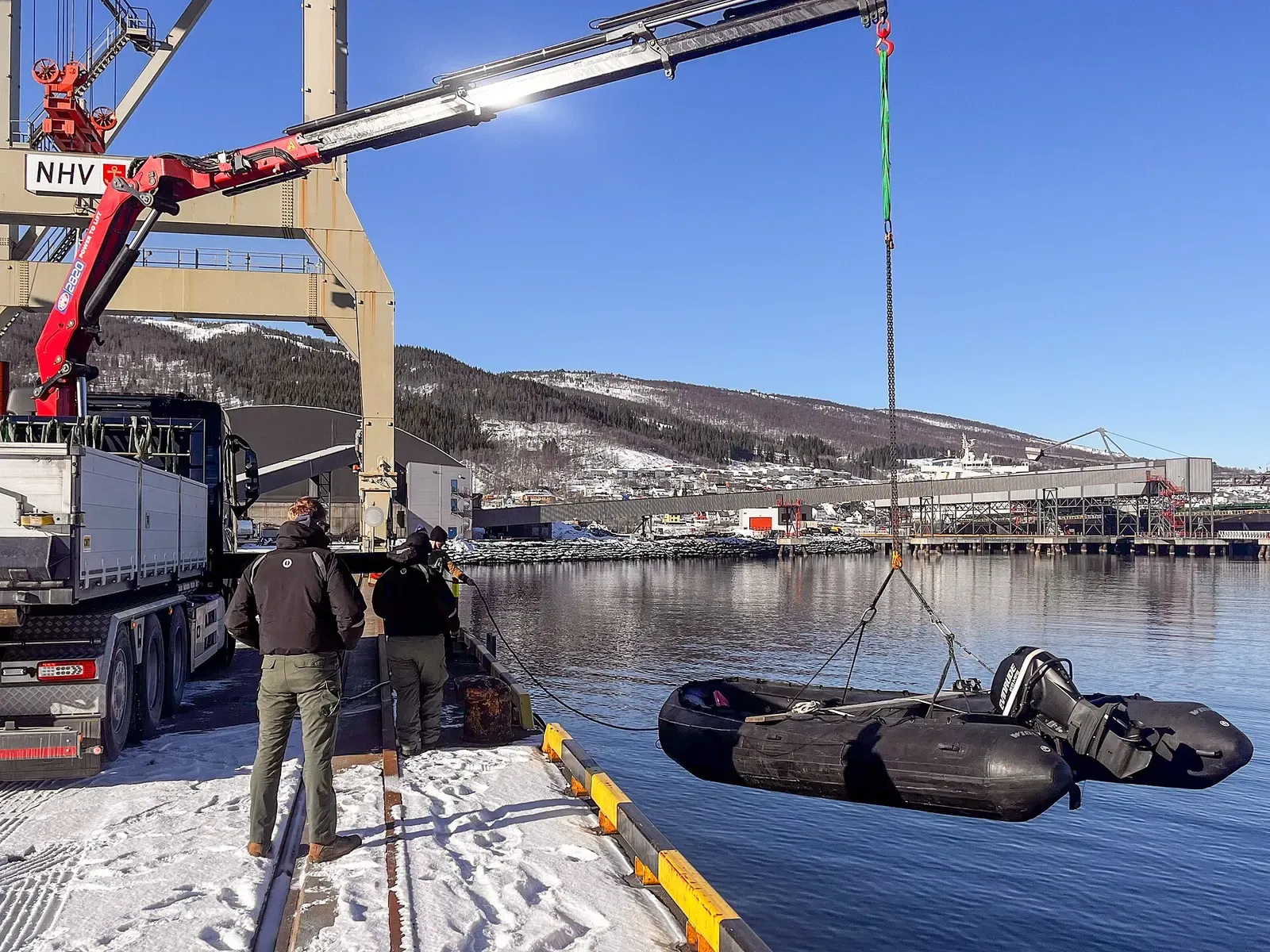 A support boat is lowered onto the water at Narvik's harbour pier, snowy peaks behind