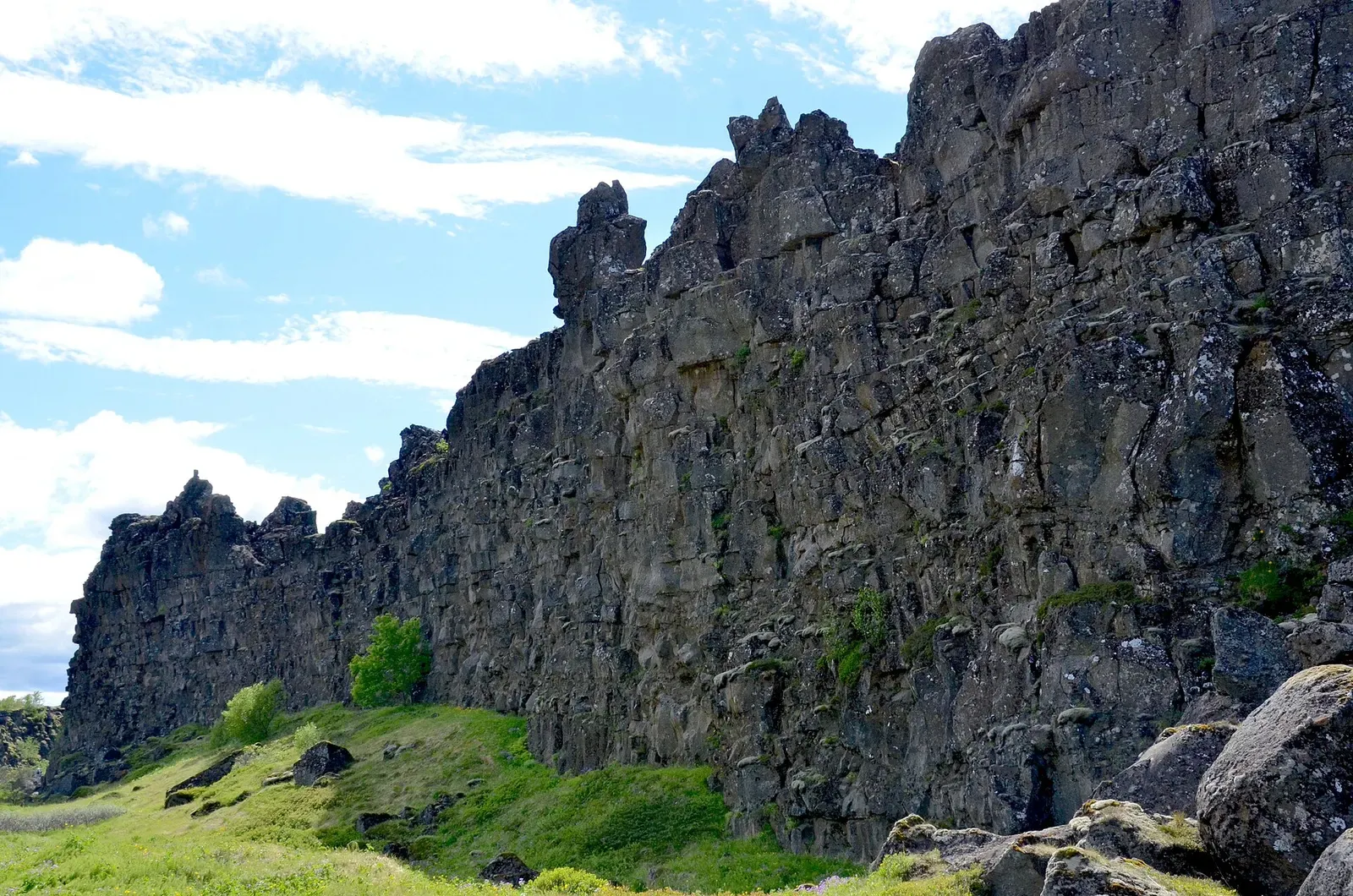 The Almannagjá rift wall rises above green grassland in Þingvellir National Park under a partly clouded sky