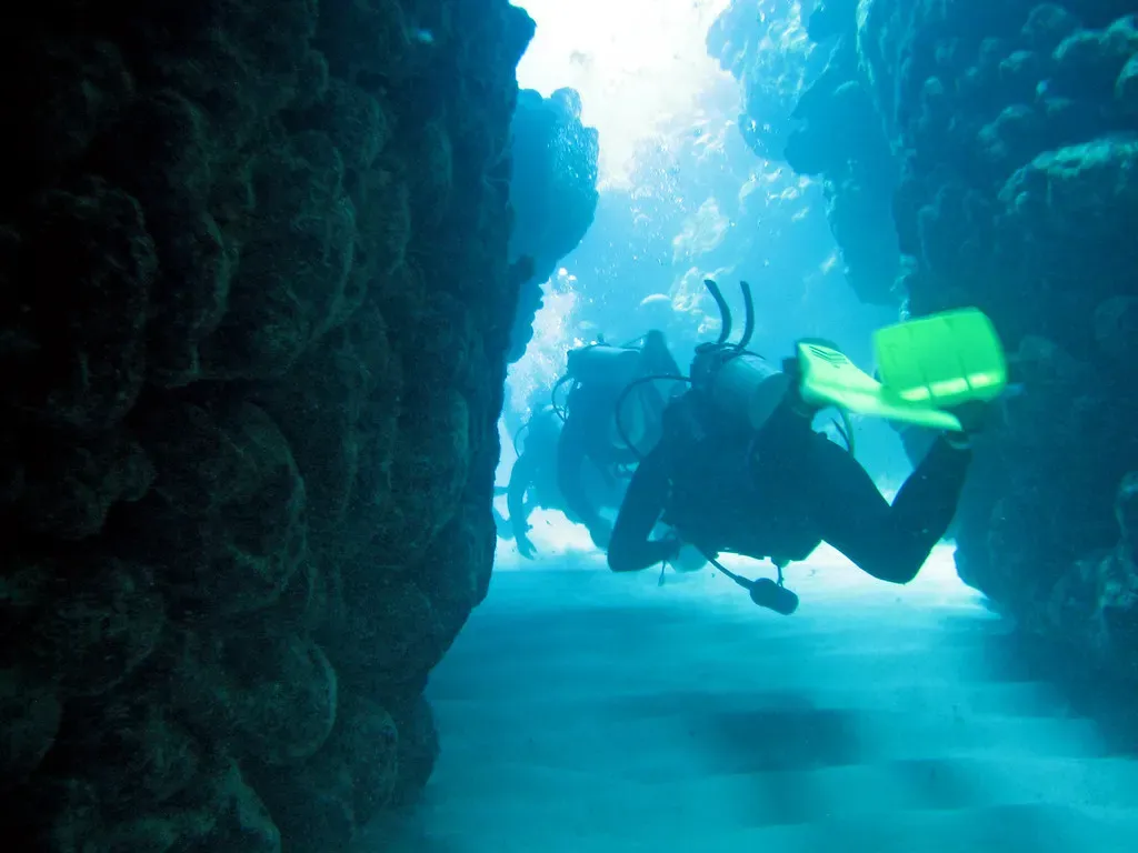 A diver with green fins swims through a narrow coral canyon lit by sunlight from above.